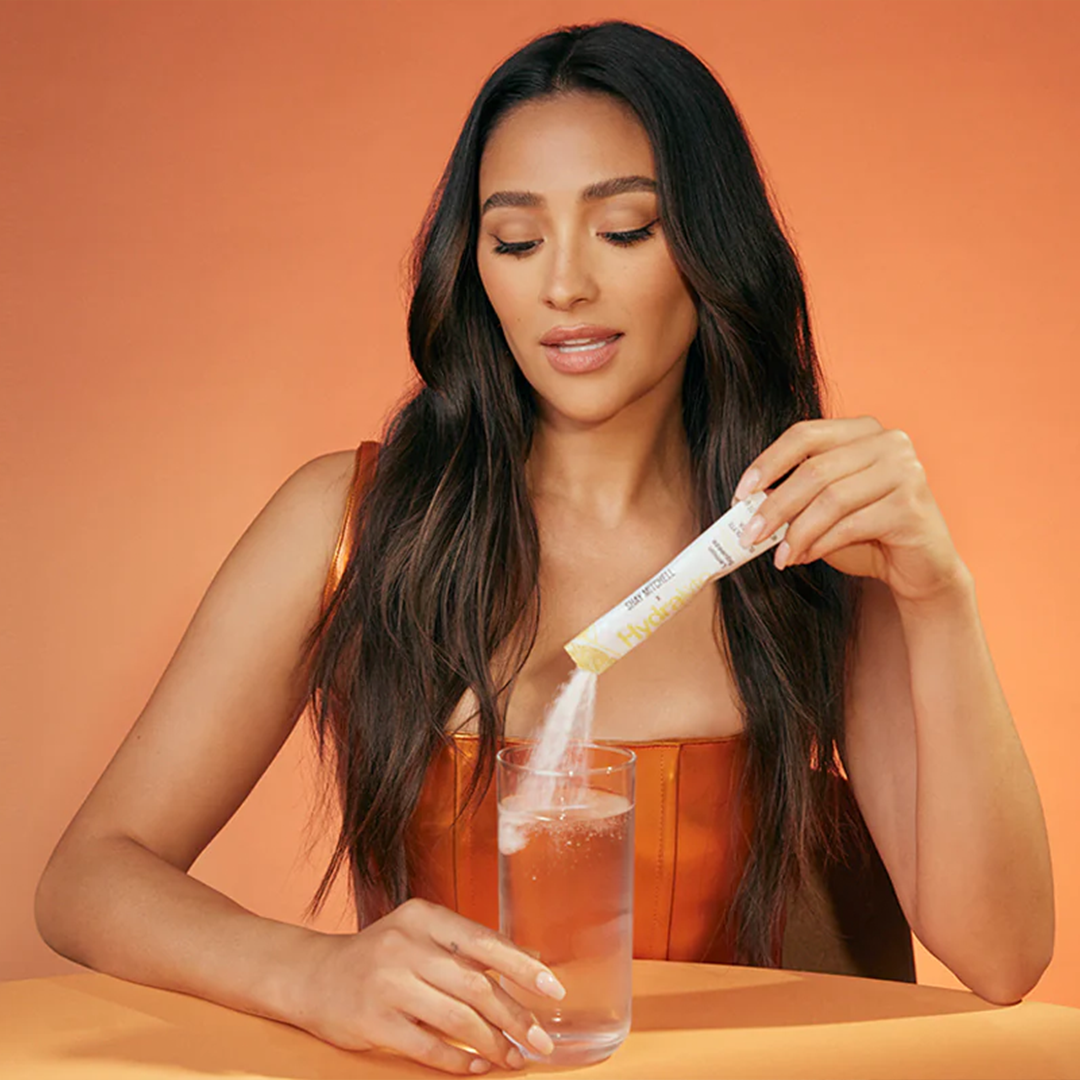 Pilar Adams Woman pours powder from a packet into a glass of water while seated at a table with an orange background. Communications & Event Strategist