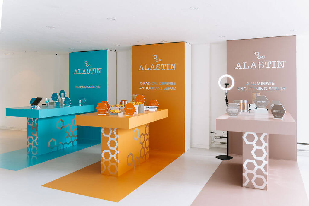 Pilar Adams A display area with three colored booths showcasing ALASTIN skincare products, each table labeled with different serums and informational plaques. Communications & Event Strategist