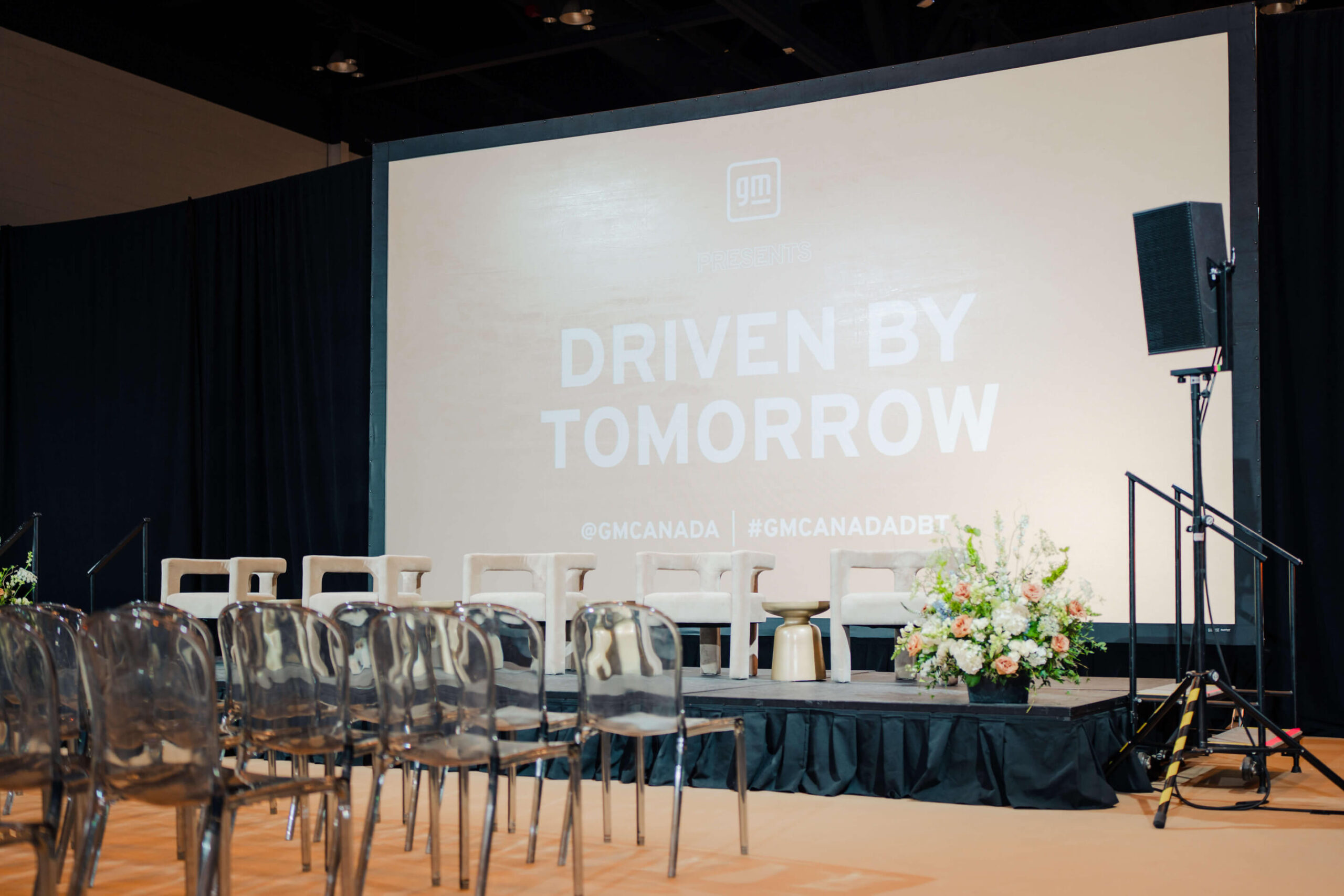 Pilar Adams A stage setup with transparent chairs, floral arrangements, and a large screen displaying "Driven by Tomorrow" at a GM Canada event. Communications & Event Strategist