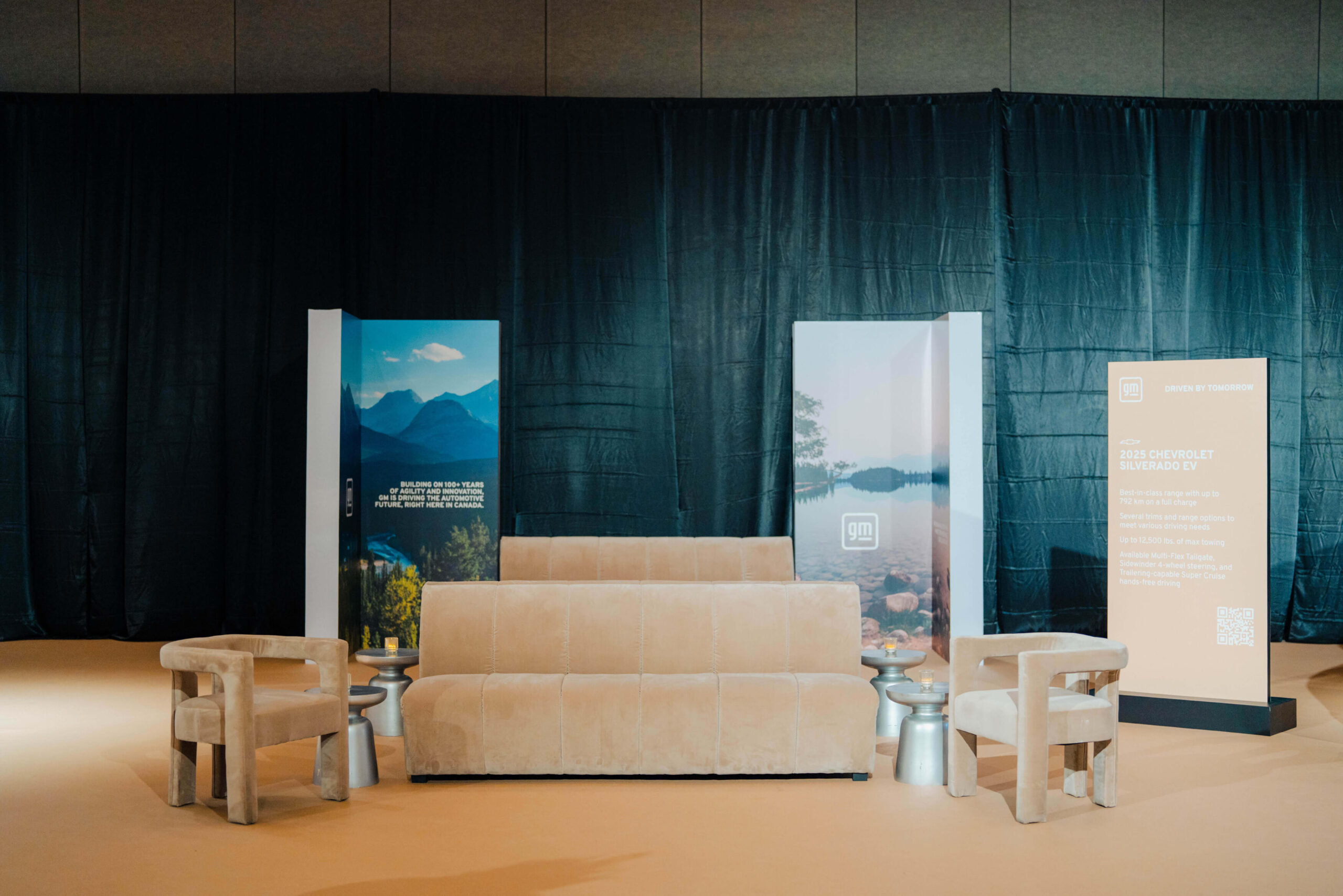 Pilar Adams A beige couch with two matching chairs and silver side tables is set in front of backdrop panels and a black curtain in a modern indoor space. Communications & Event Strategist