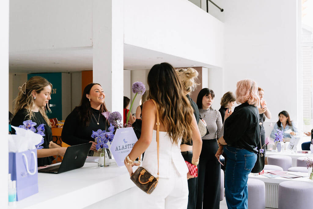 Pilar Adams People gather and interact at a modern, brightly lit reception area with a white counter and purple floral decorations. Communications & Event Strategist