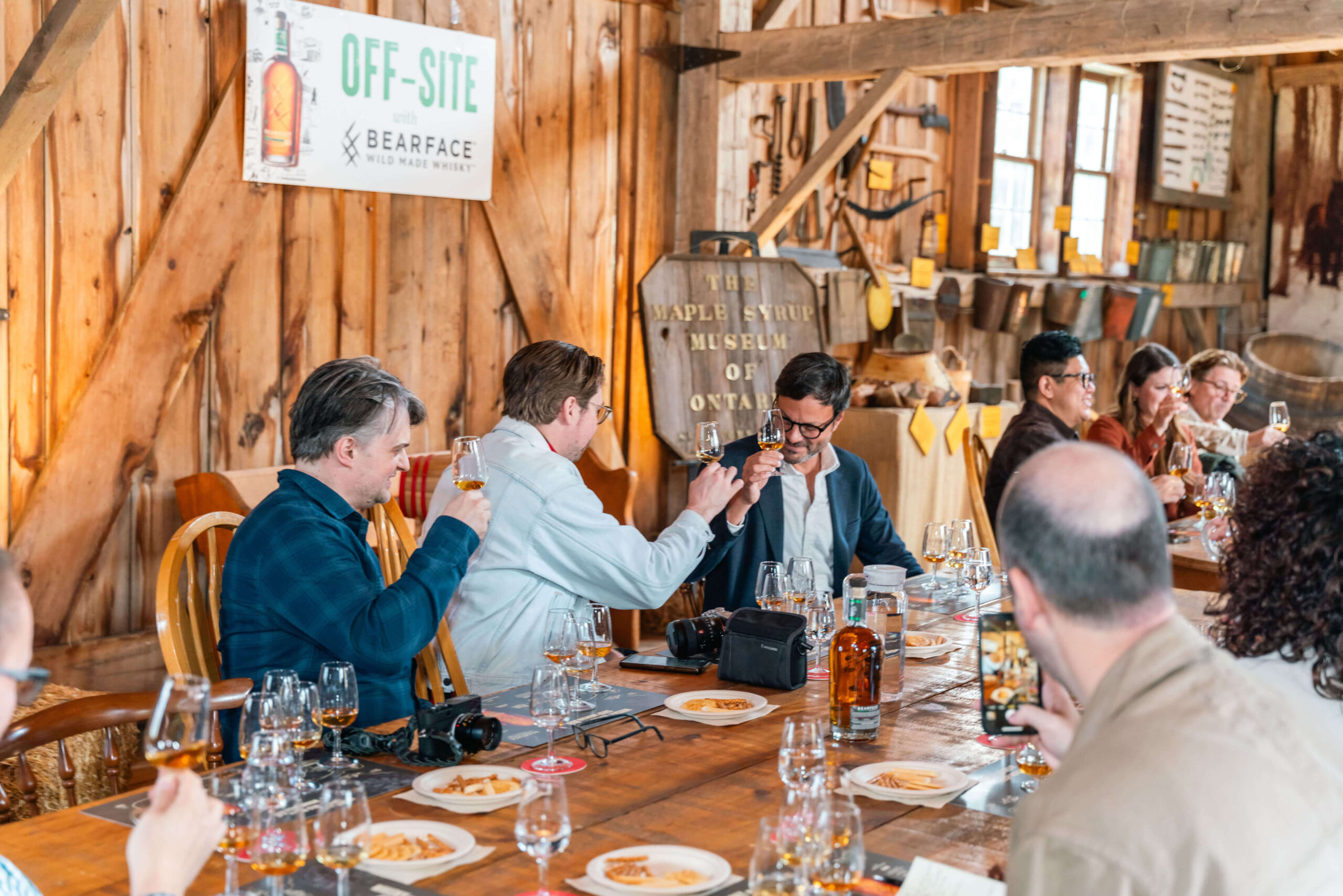 Pilar Adams A group of people sit around a wooden table in a rustic room, tasting whiskey samples from glasses with plates of food in front of them. Communications & Event Strategist
