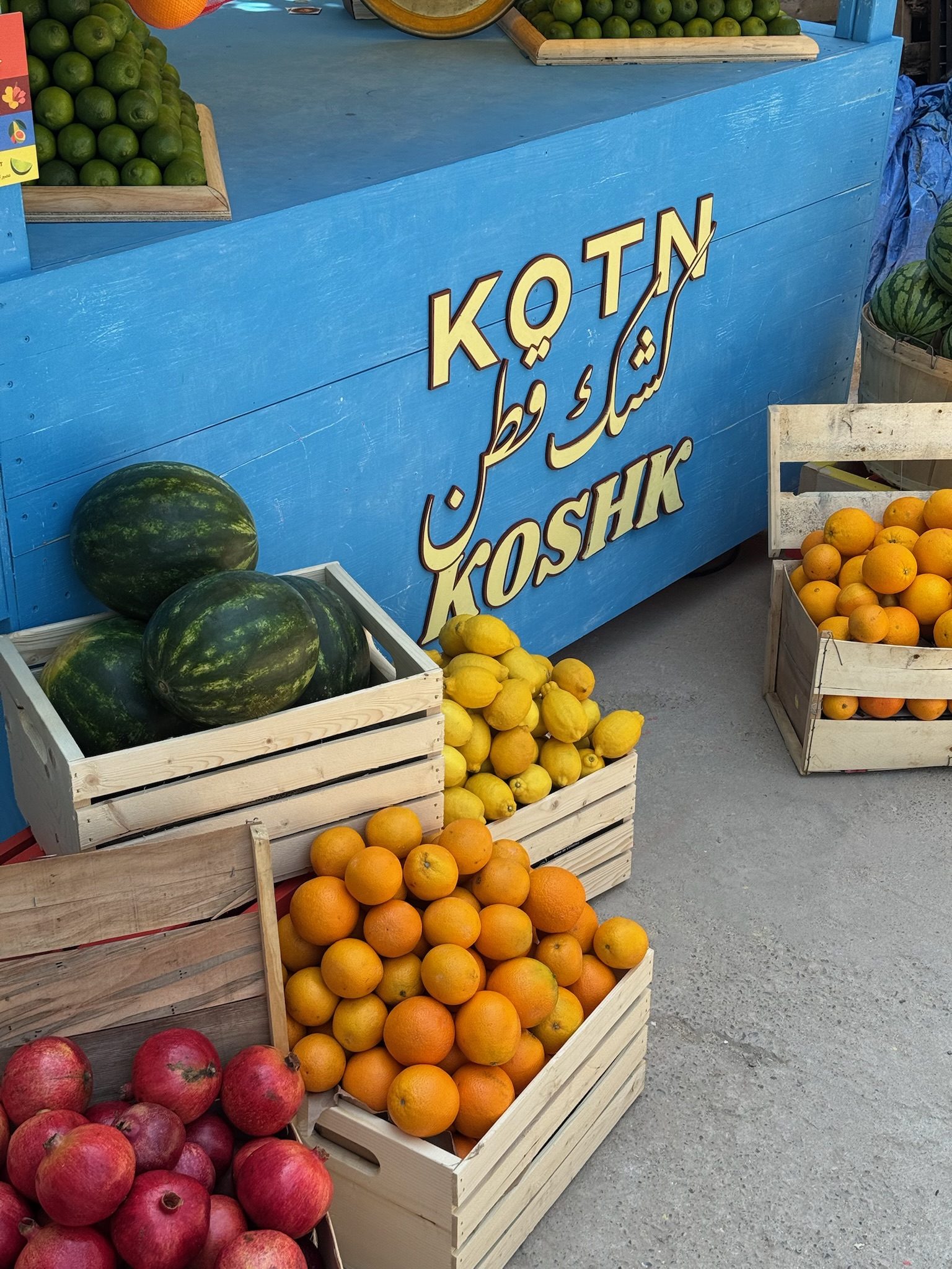 Pilar Adams Wooden crates filled with oranges, lemons, pomegranates, and watermelons are displayed in front of a blue fruit stand with "KOSH KOTN" written in English and Arabic. Communications & Event Strategist