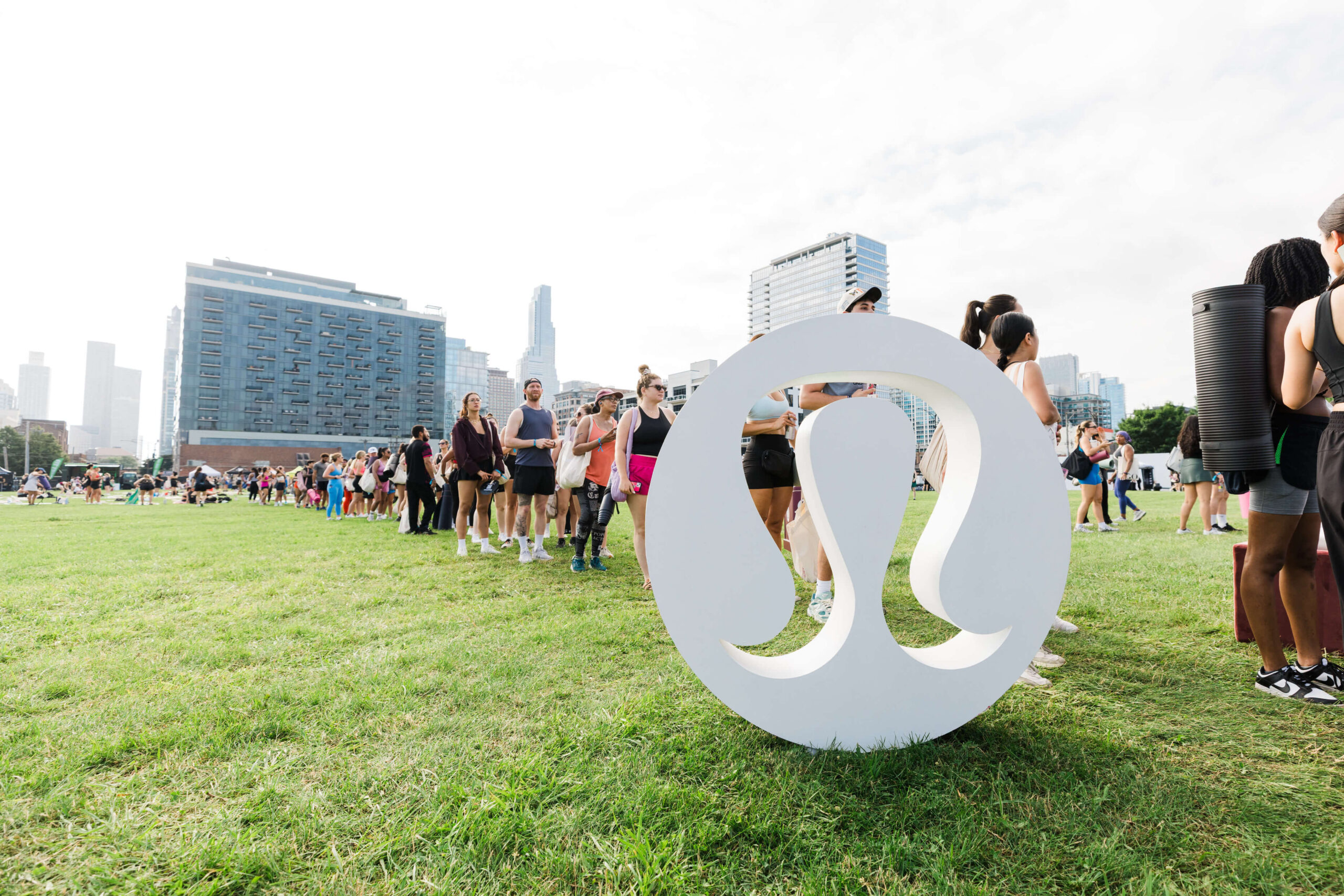 Pilar Adams People stand in line on a grassy field near a large white Lululemon logo, with city buildings in the background. Communications & Event Strategist