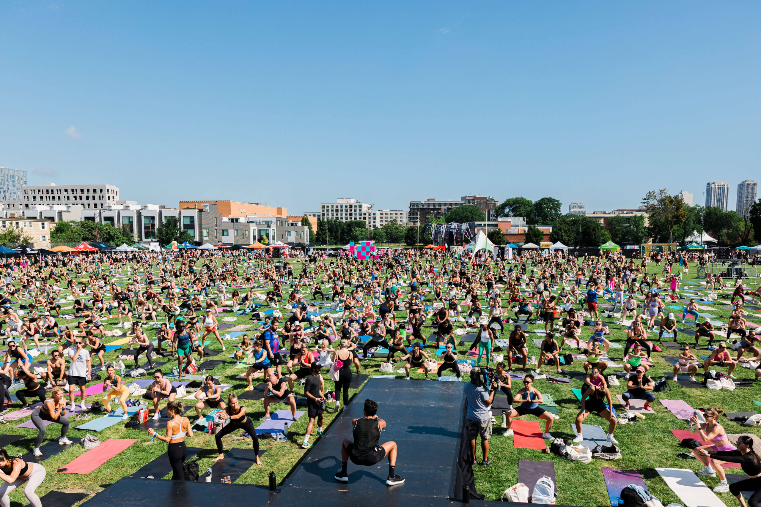 Pilar Adams A large group of people participate in an outdoor yoga class on a grassy field with a stage and instructor at the front, under a clear blue sky. Communications & Event Strategist