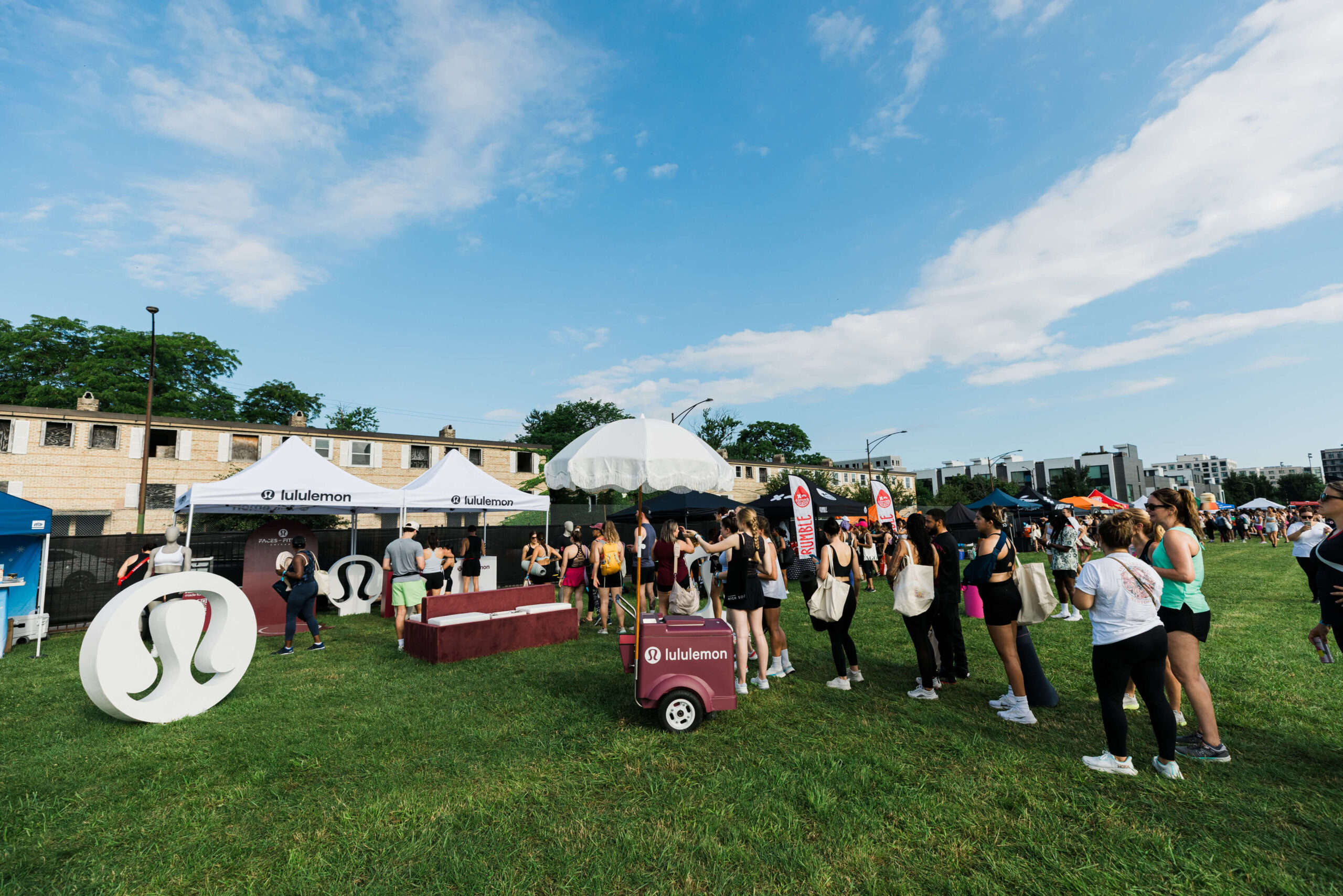 Pilar Adams A crowd gathers at a Lululemon outdoor event with branded tents, displays, and people holding tote bags on a grassy area under a partly cloudy sky. Communications & Event Strategist