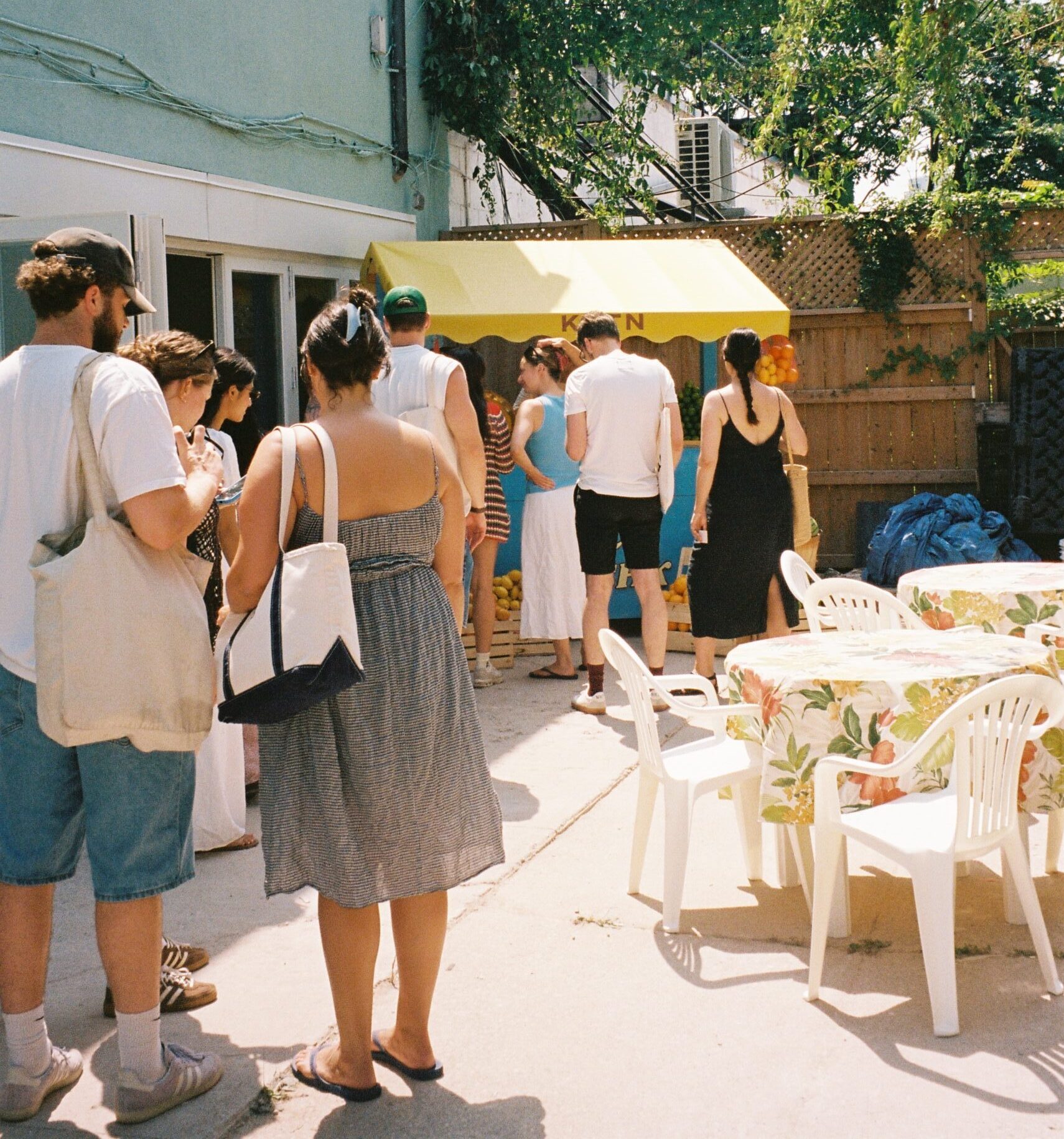 Pilar Adams A group of people stand in line outside near a yellow canopy; plastic tables and chairs with floral tablecloths are in the foreground. Communications & Event Strategist