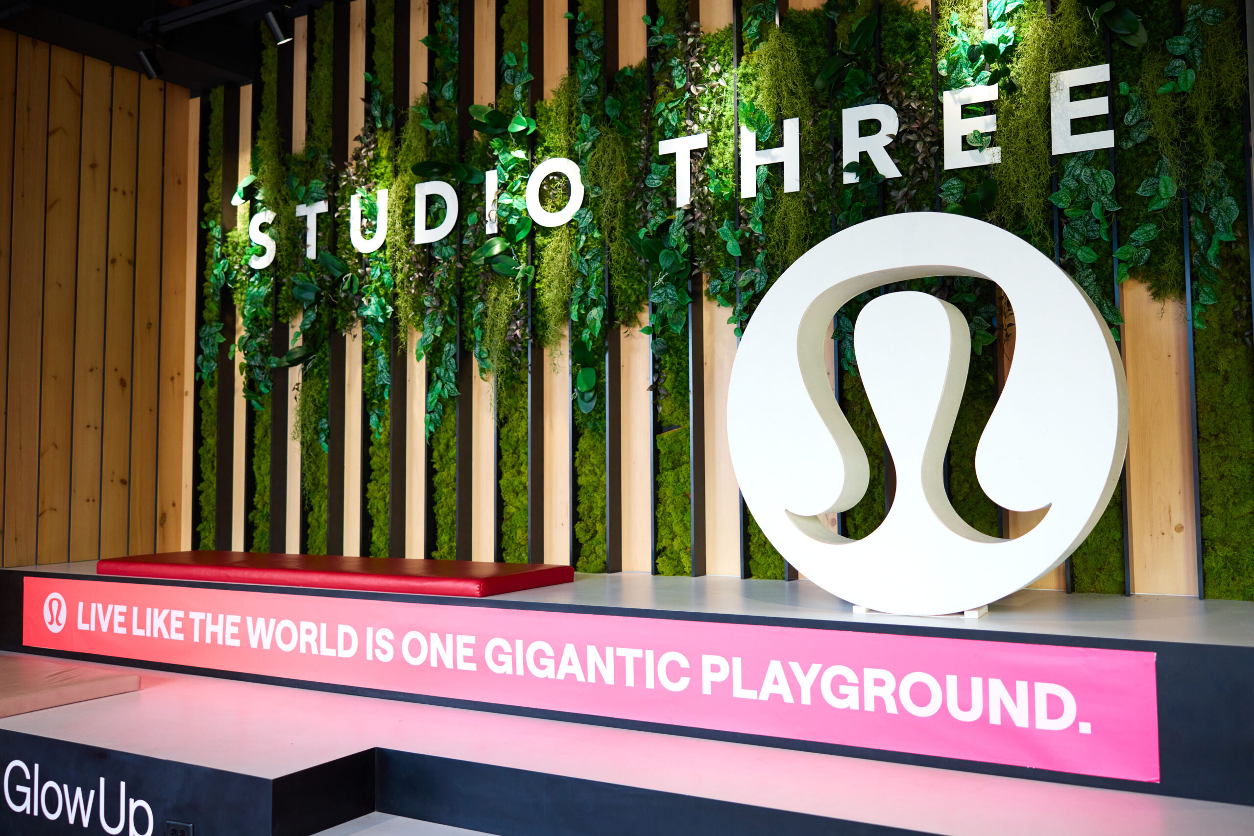 Pilar Adams Indoor stage area with "STUDIO THREE" and a large Lululemon logo, featuring greenery on the wall and a sign reading, "Live like the world is one gigantic playground. Communications & Event Strategist