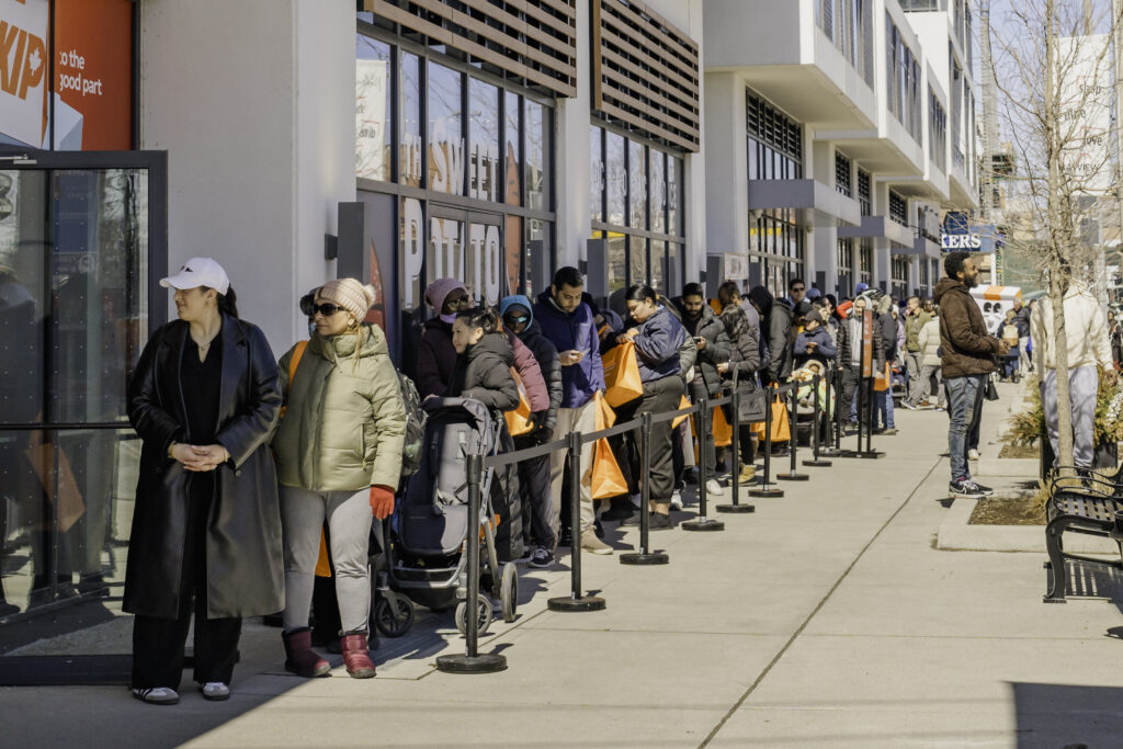 Pilar Adams A line of people wearing winter clothing stands outside a building along a sidewalk, waiting behind a barricade on a sunny day. Communications & Event Strategist